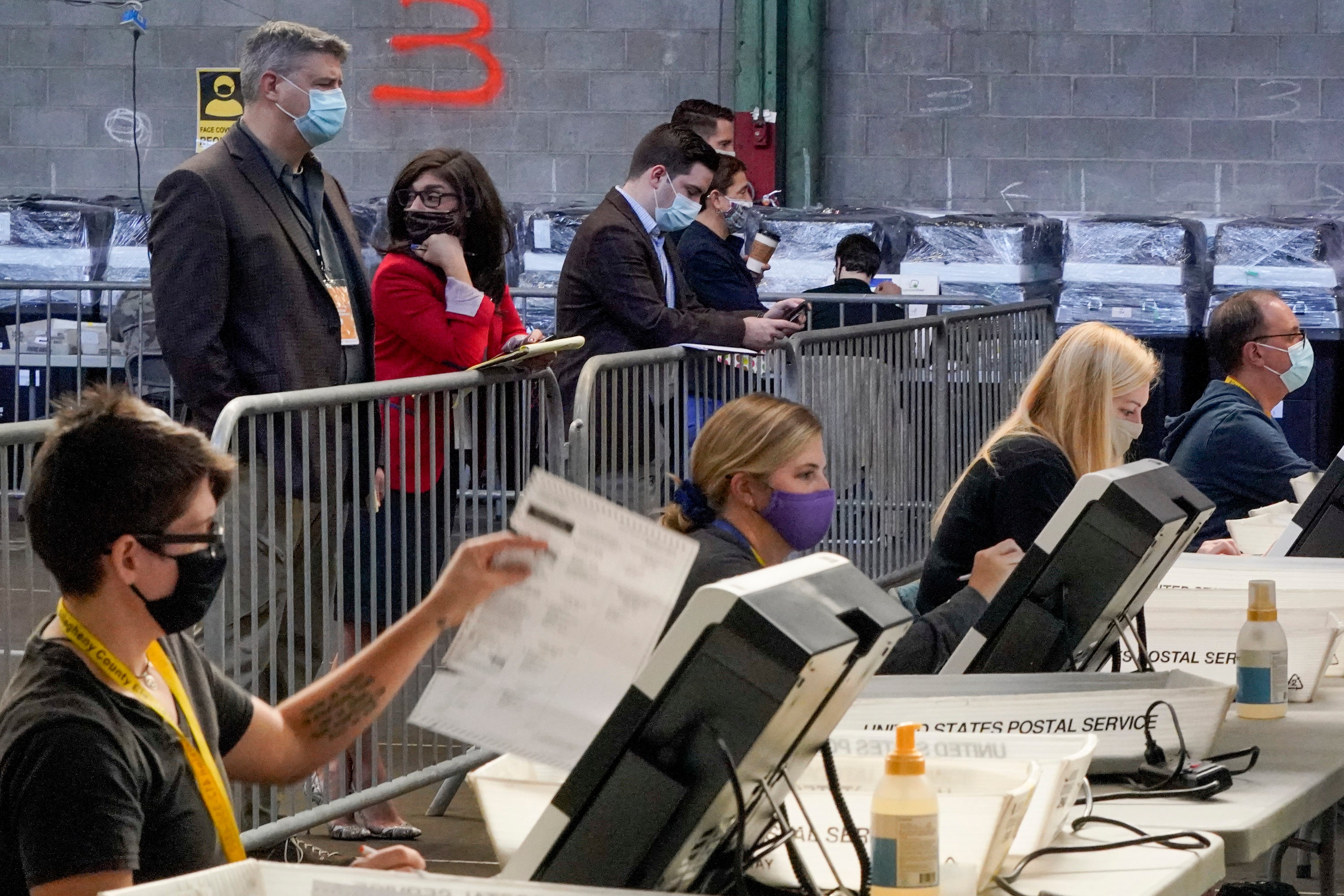 Los observadores electorales se paran detrás de una barrera y observan cómo los trabajadores de la oficina electoral procesan las boletas