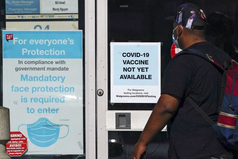 Un individuo pasa junto a una tienda de Walgreens que tiene un cartel que dice que todavia no hay disponible una vacuna contra el COVID-19 y otro que recomienda el uso de barbijos en Long Beach, California, el 2 de diciembre del 2020.