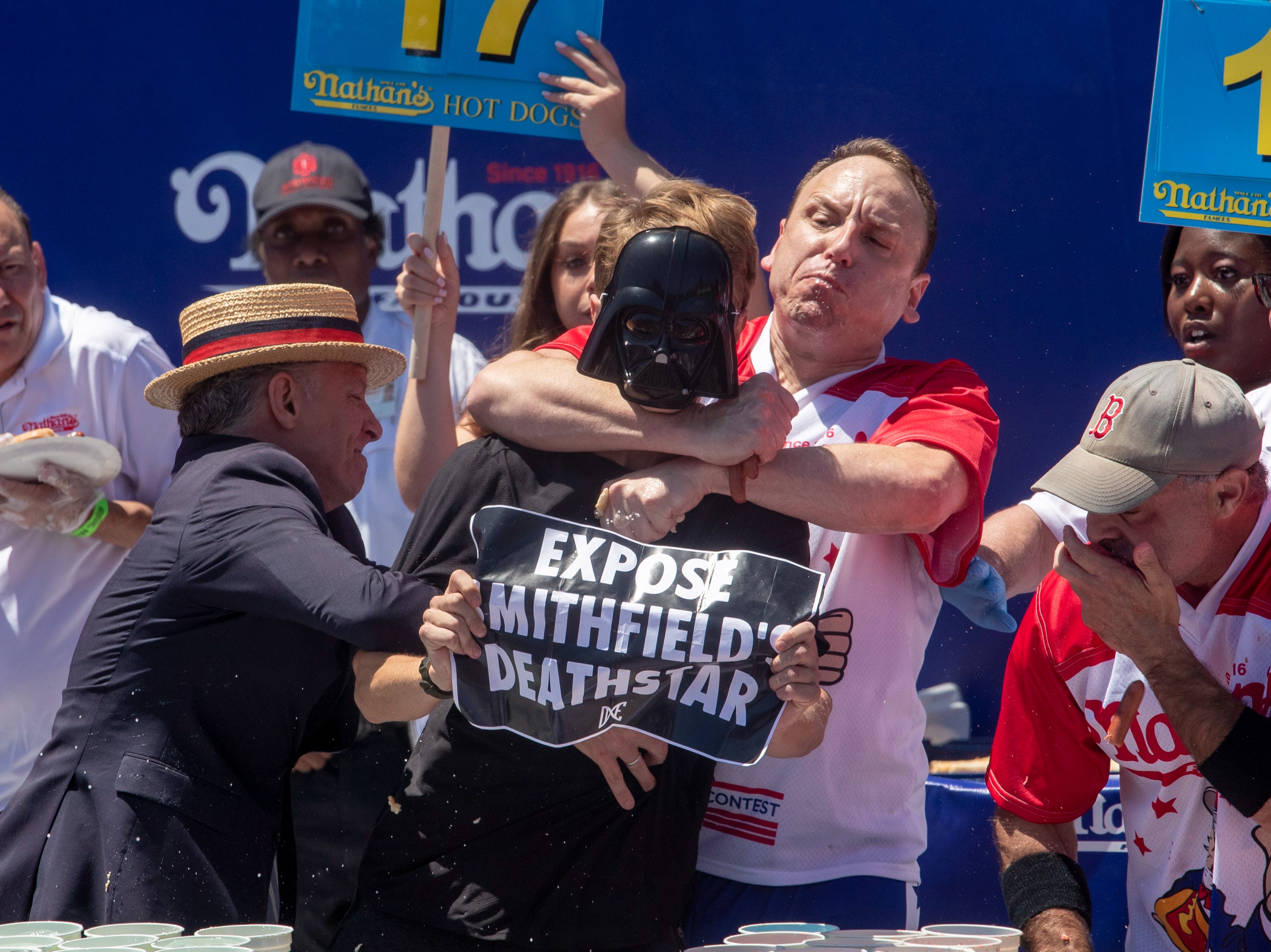 El competitivo Joey Chestnut (centro) se enfrenta a un manifestante con un cartel de “Exponer la Estrella de la Muerte de Smithfield” durante el Concurso Internacional de Hot Dogs de Nathan’s Famous del 4 de julio en Coney Island en Nueva York, Estados Unidos