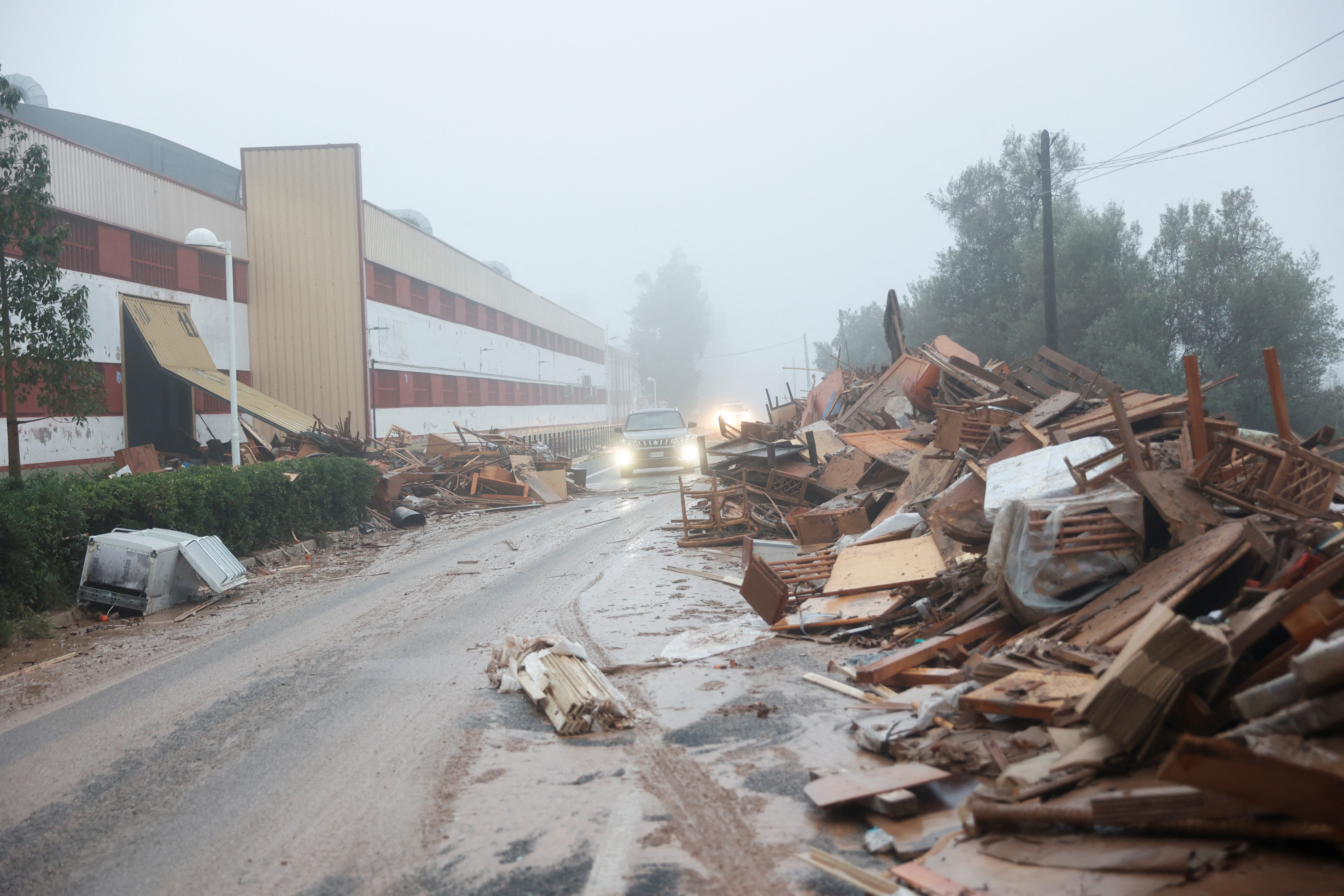 Un auto pasa junto a un montón de muebles dañados de una fábrica afectada por las inundaciones en La Alcudia (Valencia)