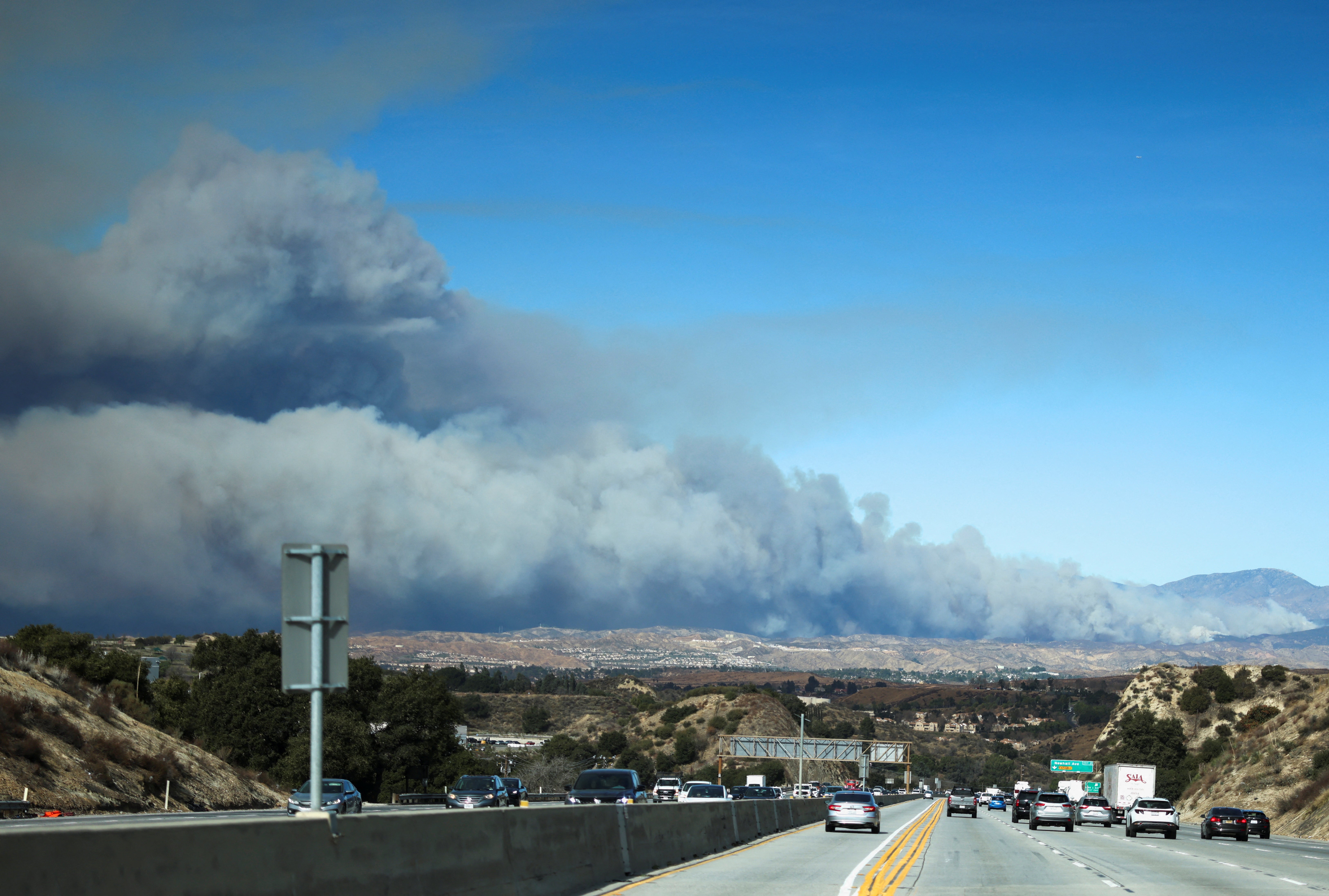 Vehículos circulan por una autopista y una nube de humo se eleva en el fondo mientras los bomberos luchan contra el incendio Hughes al norte de Santa Clarita, California, el miércoles. El incendio rápido arrasó con más de 5.000 acres en medio de un clima crítico