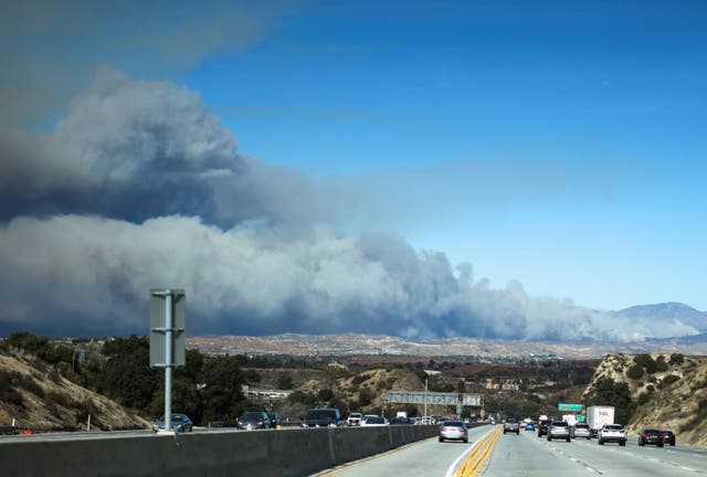 <p>Vehículos circulan por una autopista y una nube de humo se eleva en el fondo mientras los bomberos luchan contra el incendio Hughes al norte de Santa Clarita, California, el miércoles. El incendio creció rápidamente a más de 5.000 acres en medio de un clima crítico</p>