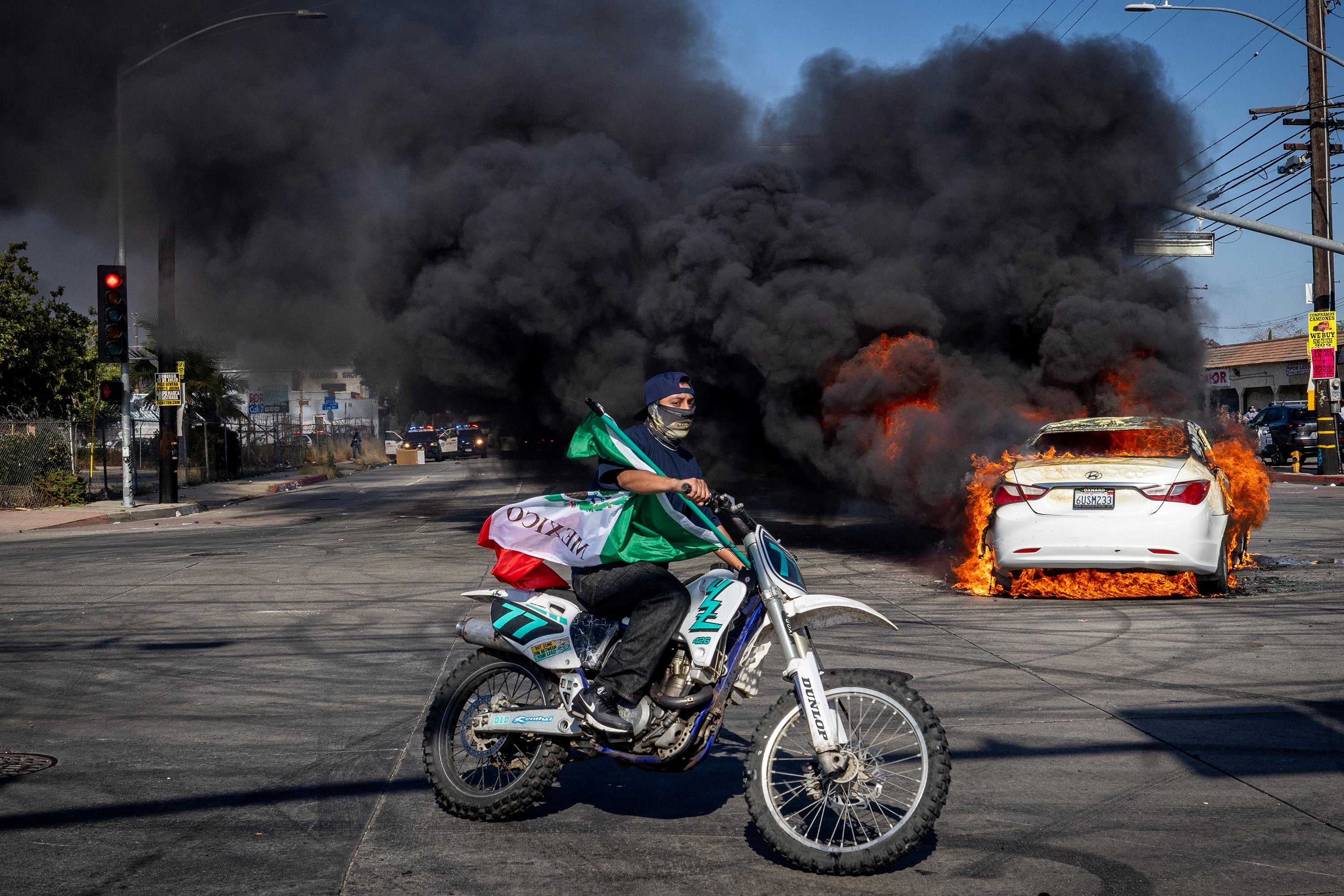 Un motociclista ondea una bandera mexicana mientras un auto en llamas arroja humo sobre Atlantic Boulevard, en medio de choques entre manifestantes y fuerzas de seguridad