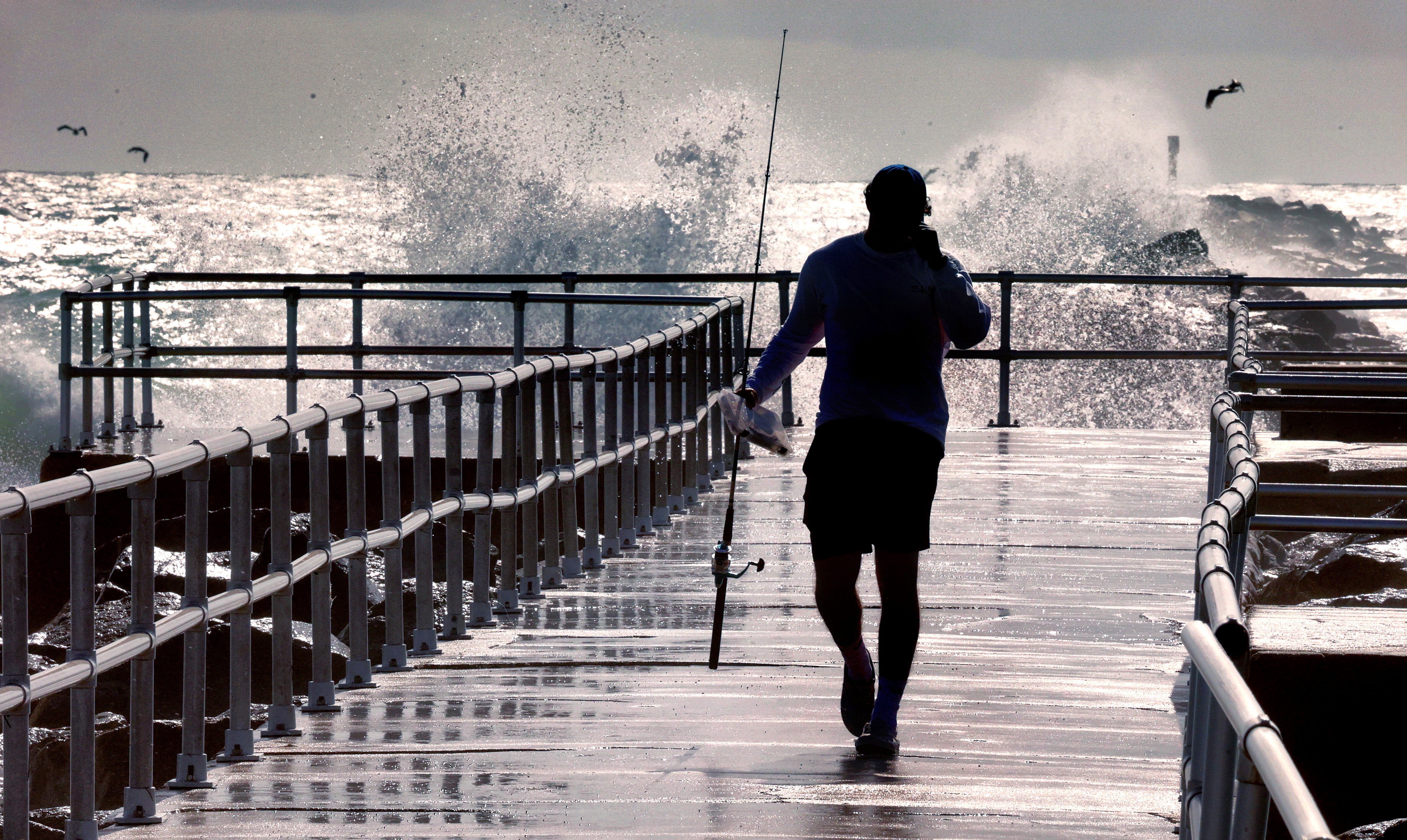 Un pescador camina por un muelle mientras las grandes olas generadas por el huracán Erin chocan contra el espigón del parque Lighthouse Point, en Ponce Inlet, Florida