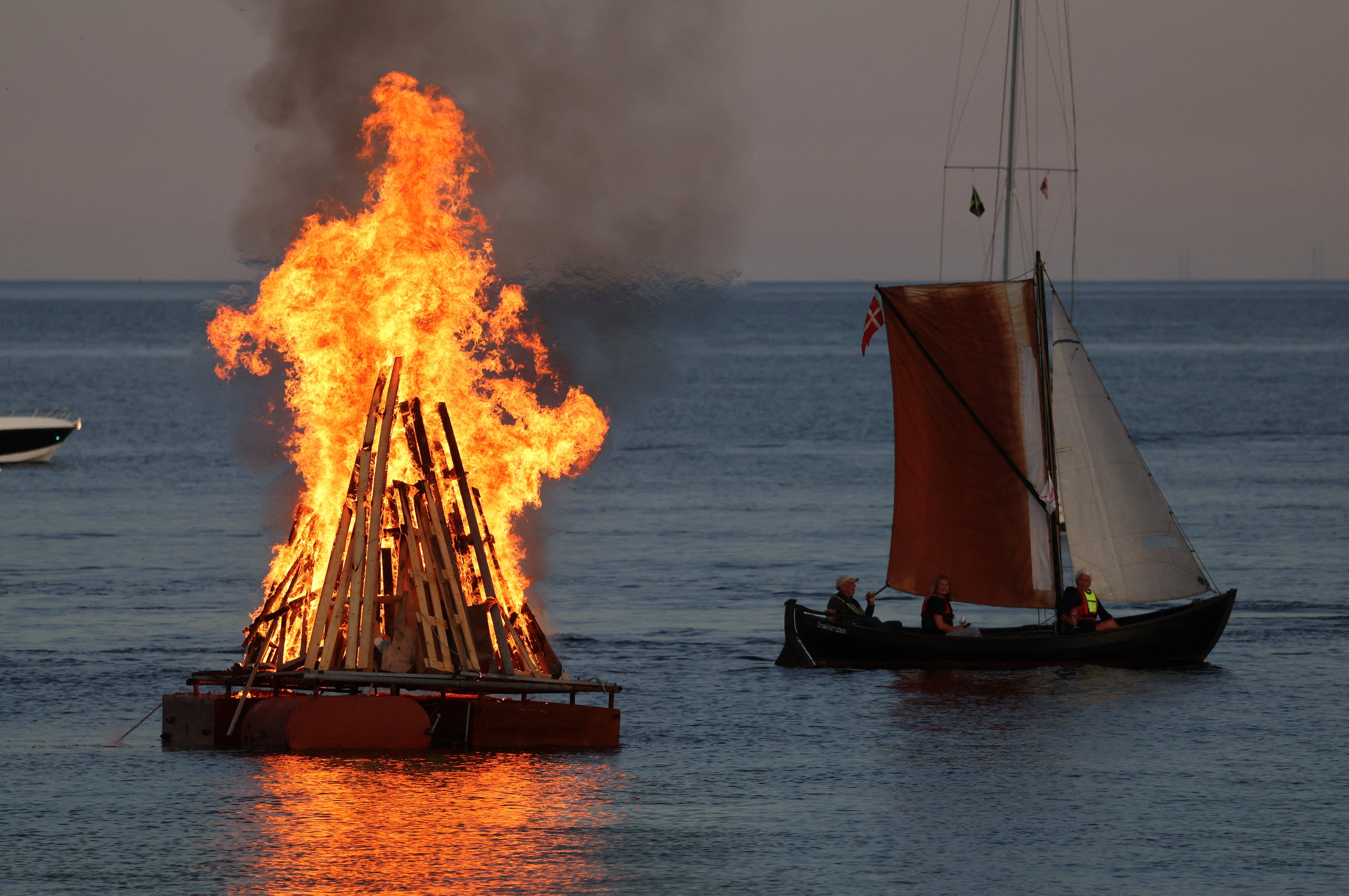 Un velero pasa junto a una hoguera del solsticio de Sankt Hans que arde en una plataforma flotante en el mar Báltico, en el puerto de Snekkersten, cerca de Helsingoer, Dinamarca, el 23 de junio de 2026. Sankt Hans combina dos tradiciones danesas: las celebraciones del solsticio de verano y la festividad cristiana de San Juan el Bautista